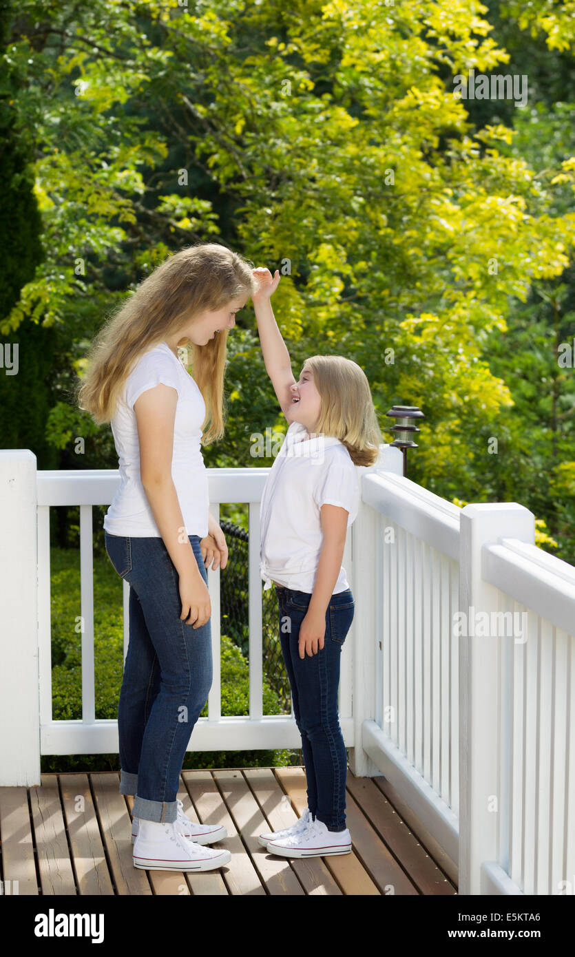 Vertikale Seitenansicht der jüngere Schwester Messung Höhenunterschied mit älteren Schwester während draußen auf der Terrasse mit unscharfen, Baum Stockfoto