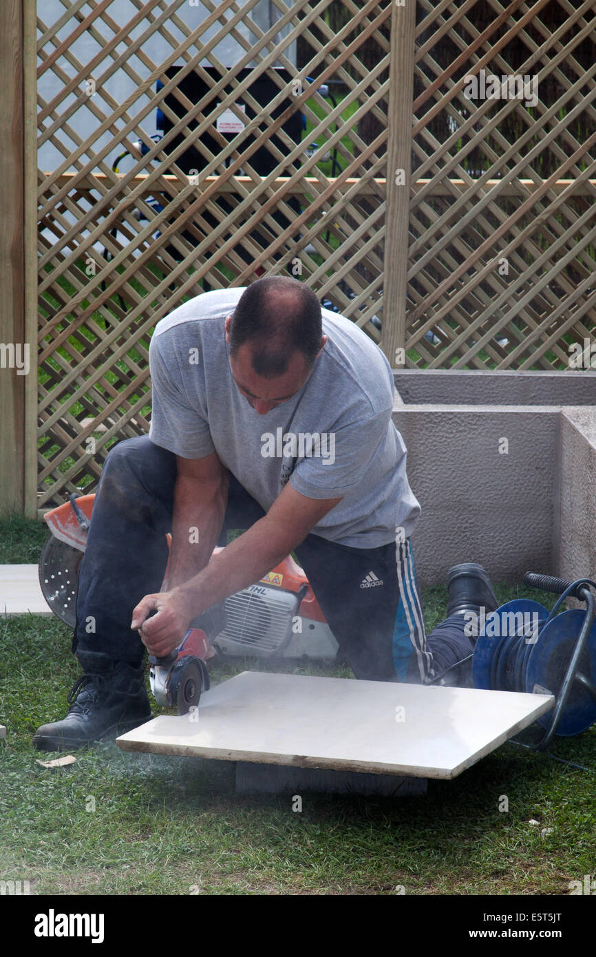 Arbeiter schneiden und legen Terrassenfliesen in Southport, Merseyside, Großbritannien. August 2014. Schneiden von Fliesen mit einem Angle Grinder als Flower Show Vorbereitungen auf den Victoria Park showgrounds, für die größte unabhängige Blumenschau Großbritanniens. Es wurde ursprünglich im Jahr 1924 von der Gemeinde gegründet, aber seit 1986 wird es von Southport Flower Show Unternehmen betrieben, die eine eingetragene Wohltätigkeitsorganisation ist. Es findet jährlich für vier Tage Ende August statt und hat eine Teilnahme von über 80.000 Personen. Stockfoto