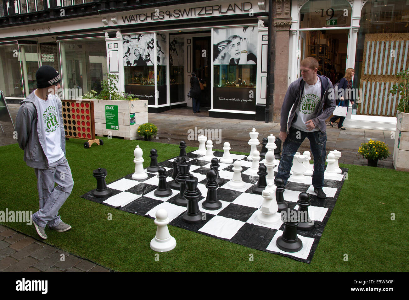 Junge Männer spielen Schach auf einem riesigen Schachbrett in der Straße,  die auf Kunstrasen verlegt ist.Manchester, Großbritannien 6th. August 2014.  Dig the City Deansgate, ein jährliches Urban Gardening Festival, das vom  Business