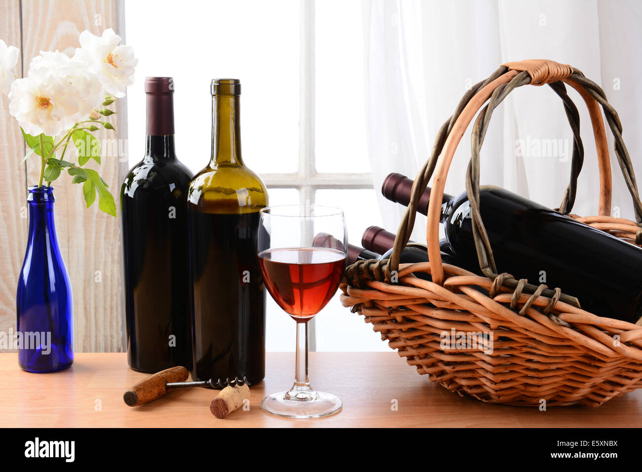 Wein Stillleben mit einem Korb voller Flaschen und einem Glas Wein und weiße Rosen in einer Vase auf einem Tisch vor einem Fenster mit Stockfoto