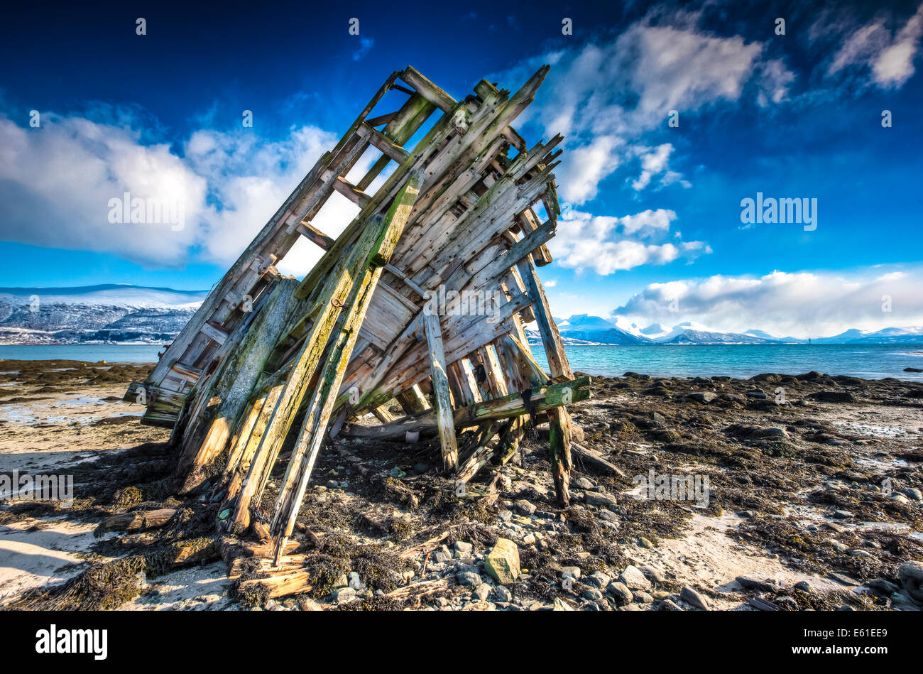 Das hölzerne Skelett eines Schiffes an einem steinigen Strand vor einem blauen bewölktem Himmel mit Schnee bedeckten Berge hinter zerstört Stockfoto