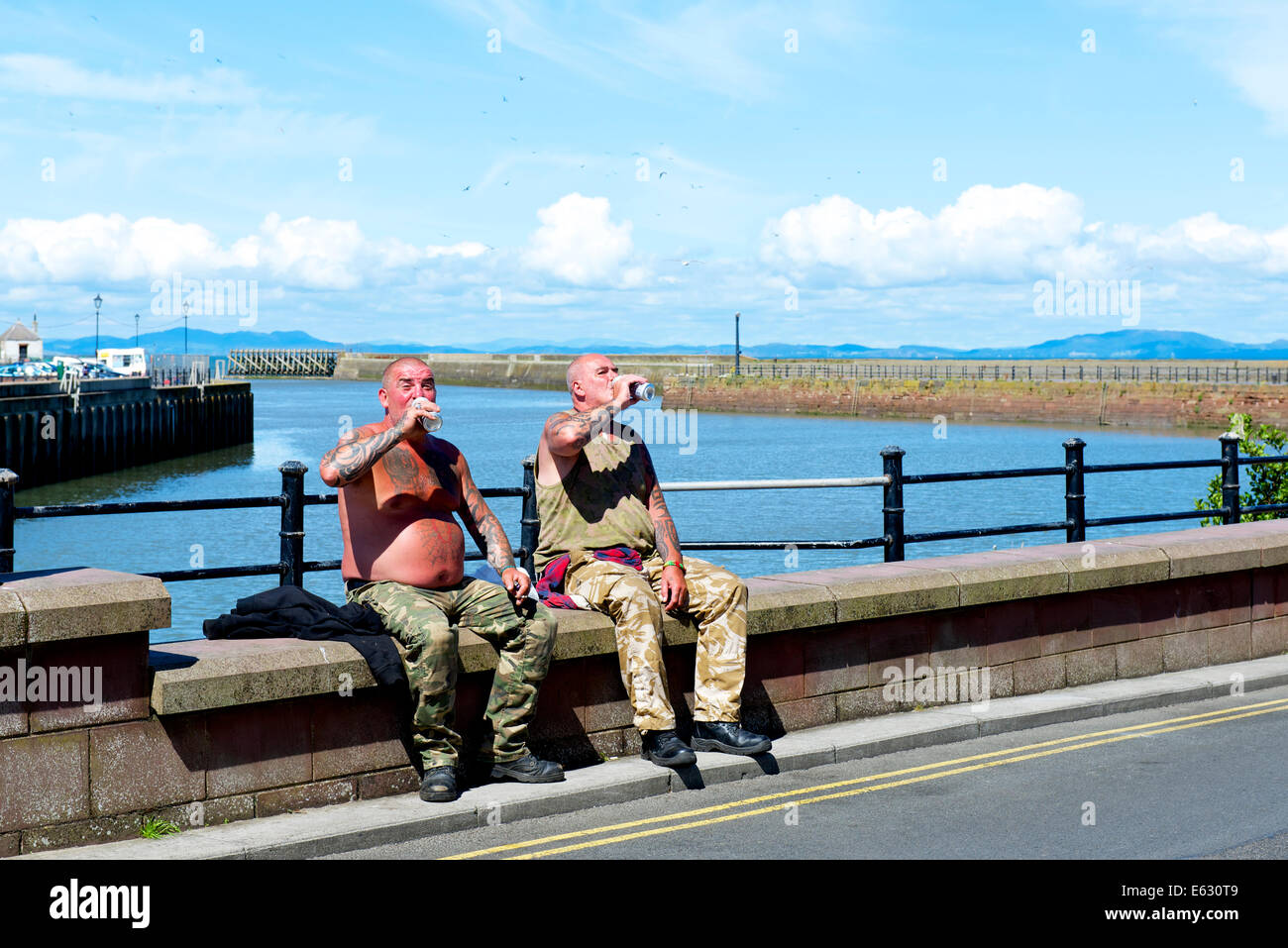 Zwei mittlere Alter, tätowierte Männer, sitzen auf der Hafenmauer, trinken Bier aus Dosen, in Maryport, West Cumbria, England UK Stockfoto