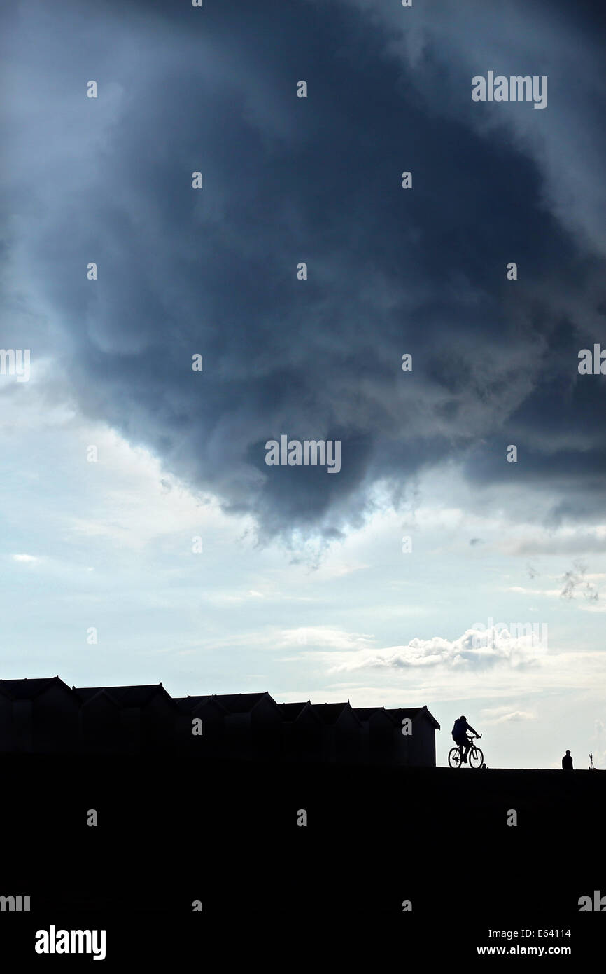 Gewitterwolken Brauen über Strandhütten und Boote an der Strandpromenade von Göring, West Sussex, wie Radfahrer und Menschen vorbeigehen Stockfoto