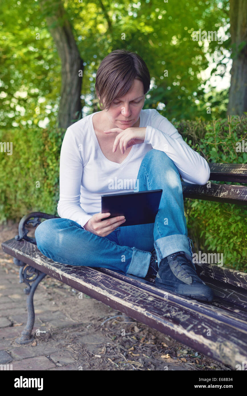 Frau liest E-Book auf digitale Tablettcomputer sitzend auf einer Holzbank im Park. Stockfoto