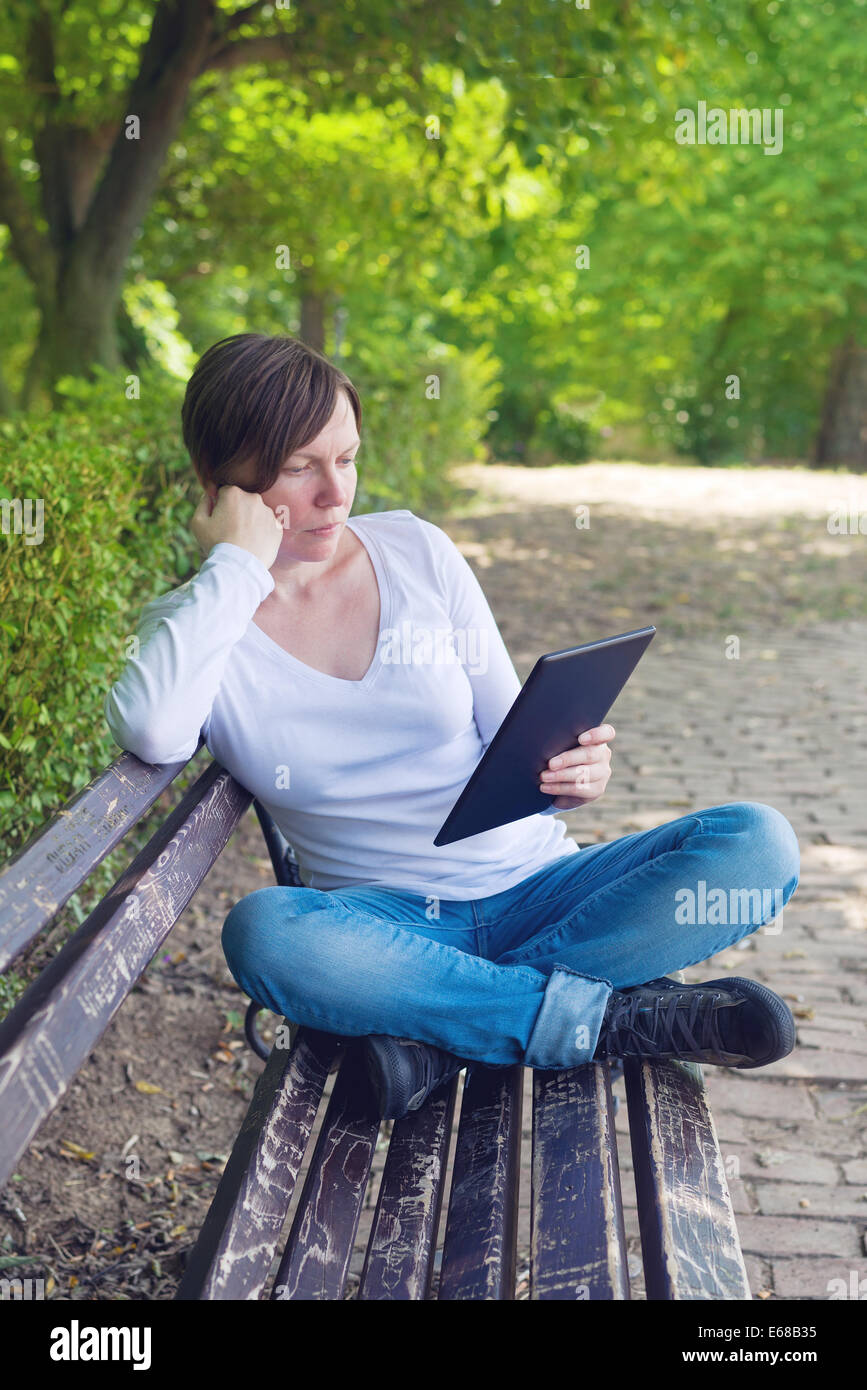 Frau liest E-Book auf digitale Tablettcomputer sitzend auf einer Holzbank im Park. Stockfoto