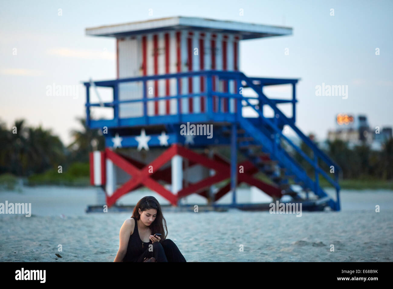 Eine Dame saß am South Beach in Miami SMS mit Start und Streifen Strandhütte hinter Ihnen Stockfoto