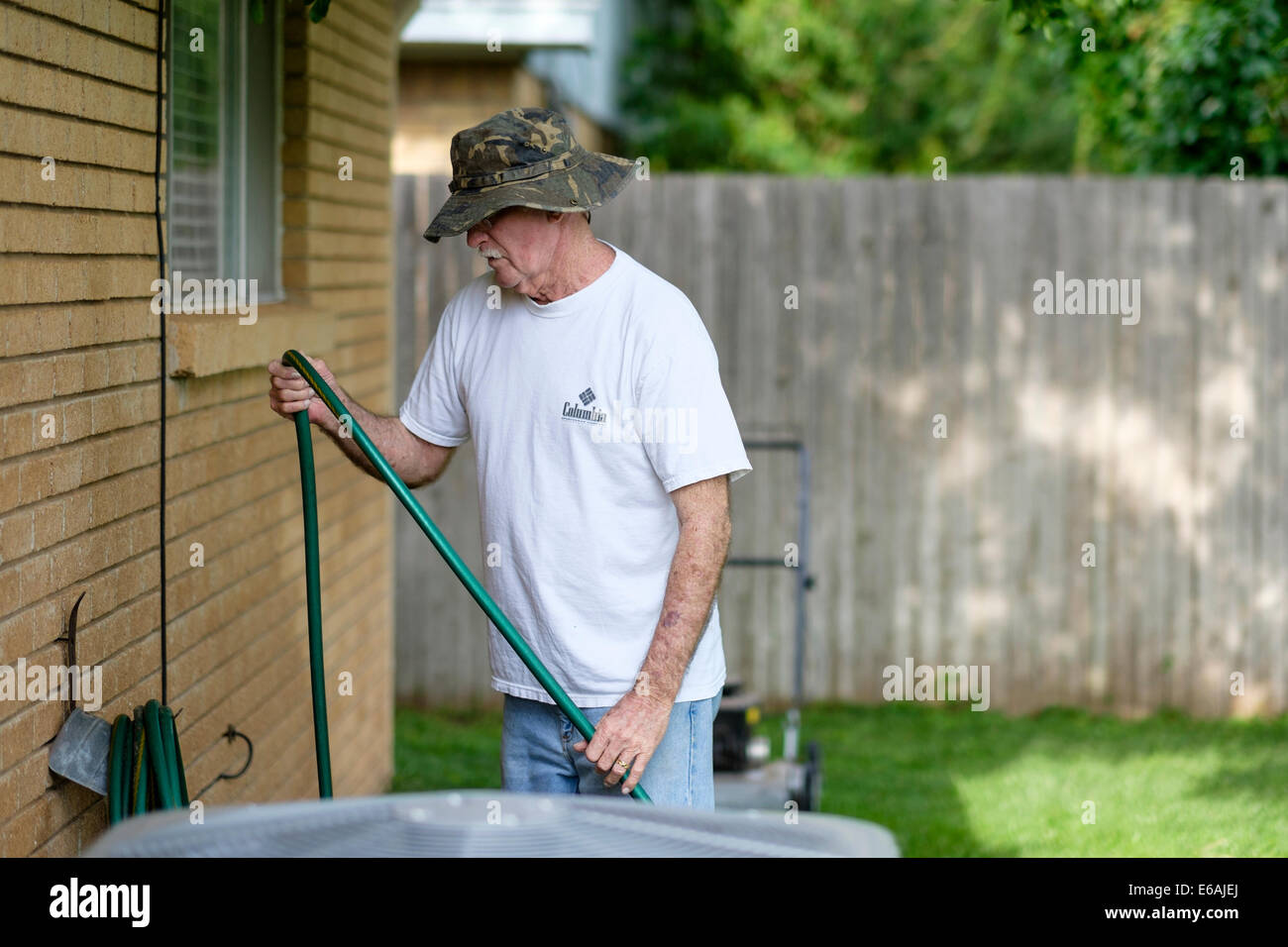 Ein 76-jähriger Kaukasier erledigt Hofarbeiten, indem er draußen einen Wasserschlauch gegen das Haus wickelt. USA Stockfoto