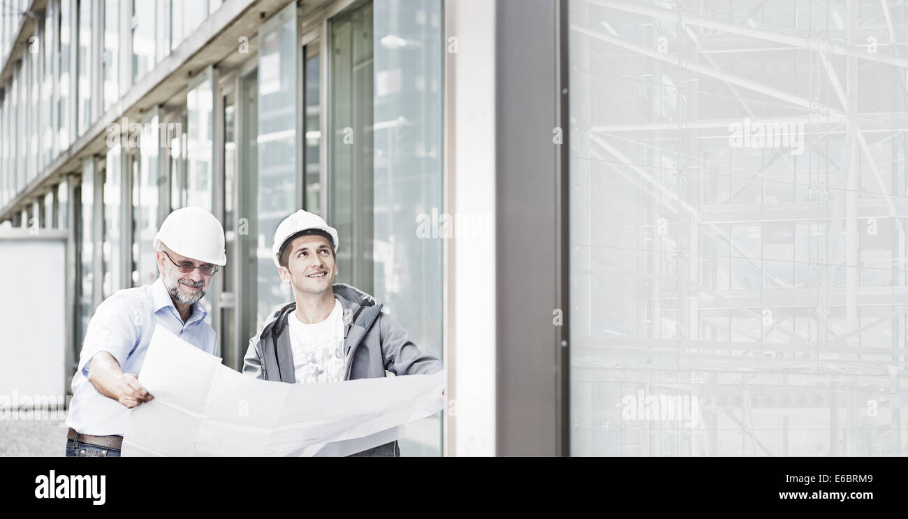 Zwei Ingenieure, 55-65 und 25-35 Jahre alt, planen harte Hüte tragen, stehen vor einem Gebäude und Blick auf ein Gebäude Stockfoto