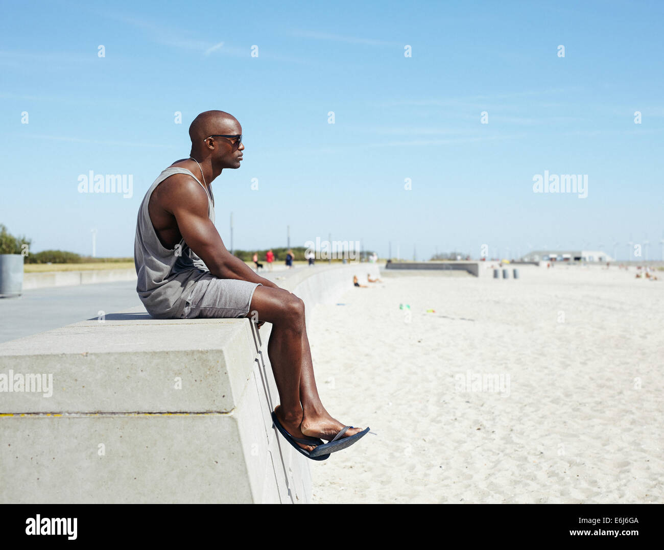 Seitenansicht der jungen afrikanischen Mann sitzt auf einer Strandpromenade wegsehen. Afro amerikanische Modell entspannen im Freien am Strand mit co Stockfoto