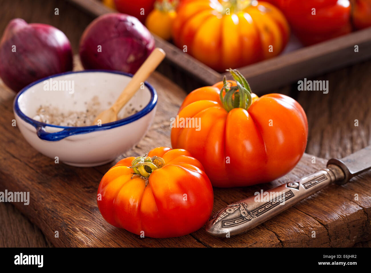Reife Fleischtomaten aus dem eigenen Garten mit einem Messer, Zwiebeln und Salz auf ein altes rustikales Schneidebrett im Landhausstil Stockfoto