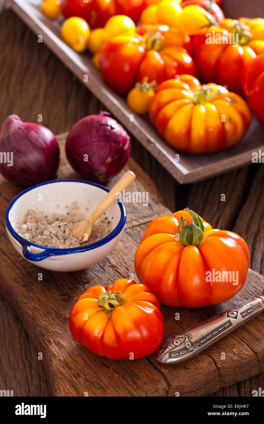 Reife Fleischtomaten aus dem eigenen Garten mit einem Messer, Zwiebeln und Salz auf ein altes rustikales Schneidebrett im Landhausstil Stockfoto