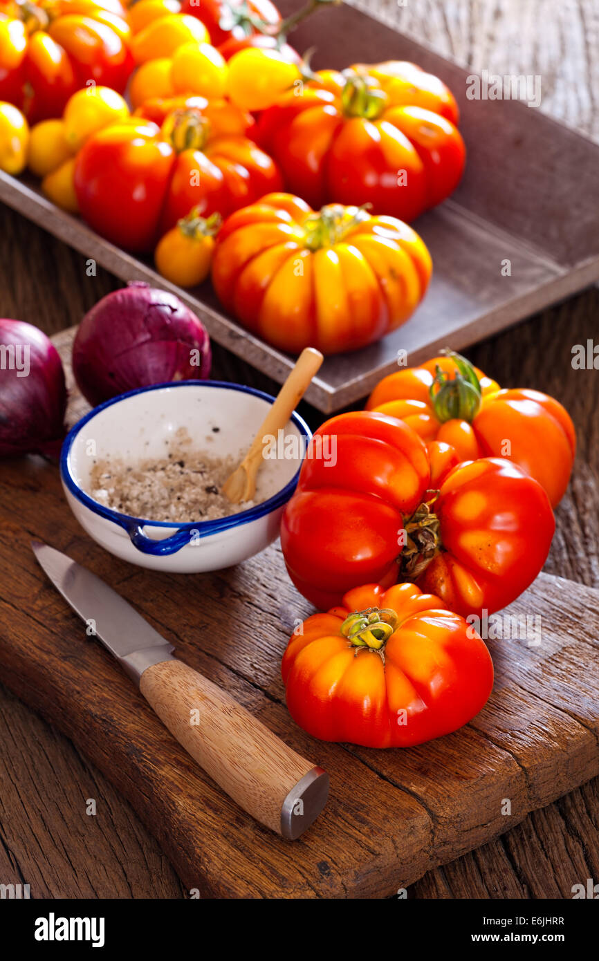 Reife Fleischtomaten aus dem eigenen Garten mit einem Messer, Zwiebeln und Salzstreuer auf einem alten rustikalen Schneidebrett im Landhausstil Stockfoto