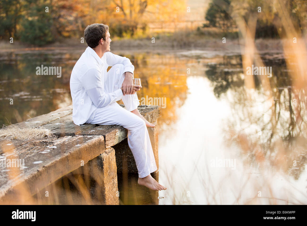 junger Mann sitzen auf Pier mit Wein und Blick auf den Sonnenuntergang am See genießen Stockfoto