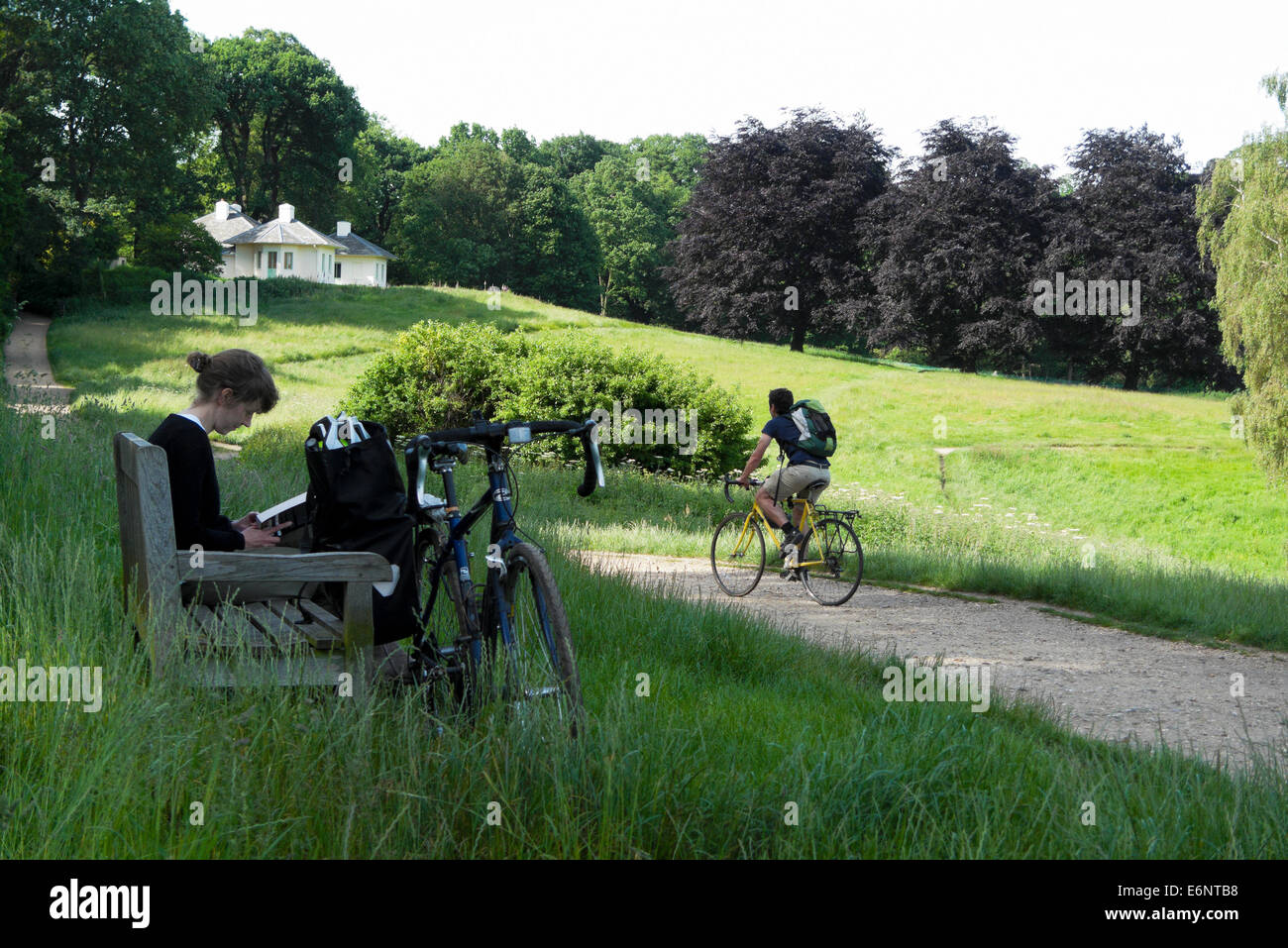 Radfahrer fährt Fahrrad vorbei an eine Frau liest ein Buch auf einer Bank sitzen, im Sommer auf Hampstead Heath North London NW3 UK KATHY DEWITT Stockfoto