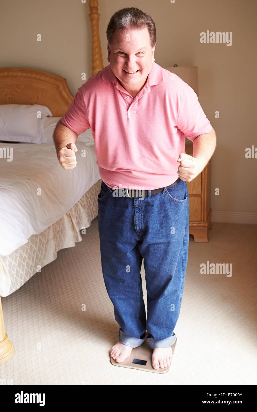 Glücklich übergewichtiger Mann stehend auf Skalen im Schlafzimmer Stockfoto
