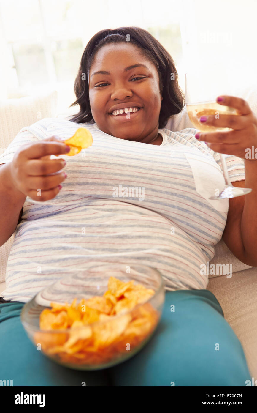 Übergewichtige Frau zu Hause essen Chips und trinken Wein Stockfoto