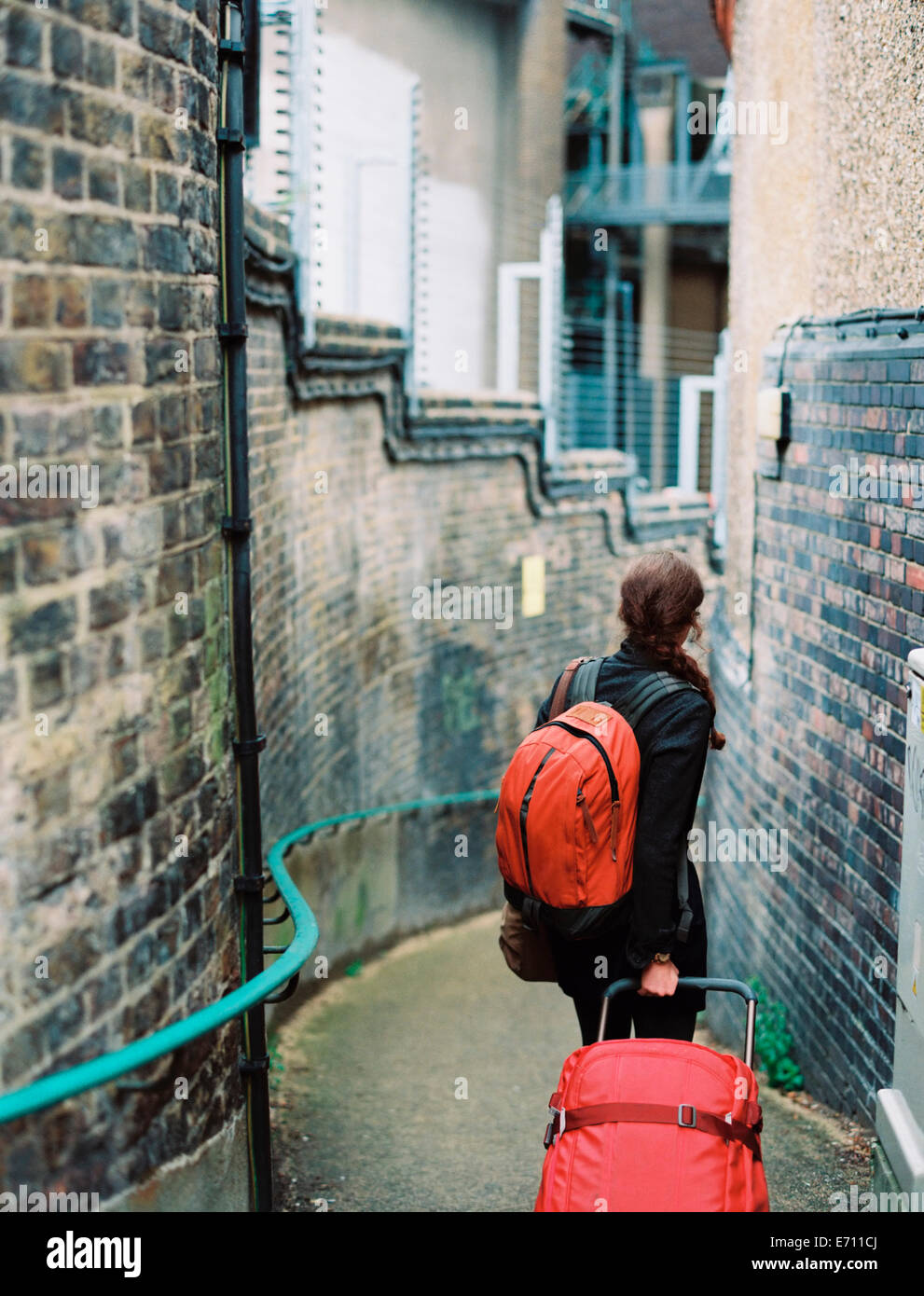 Eine Frau Mauerwerk in einer schmalen Straße, ziehen einen Koffer und einen orangenen Rucksack holdin. Stockfoto