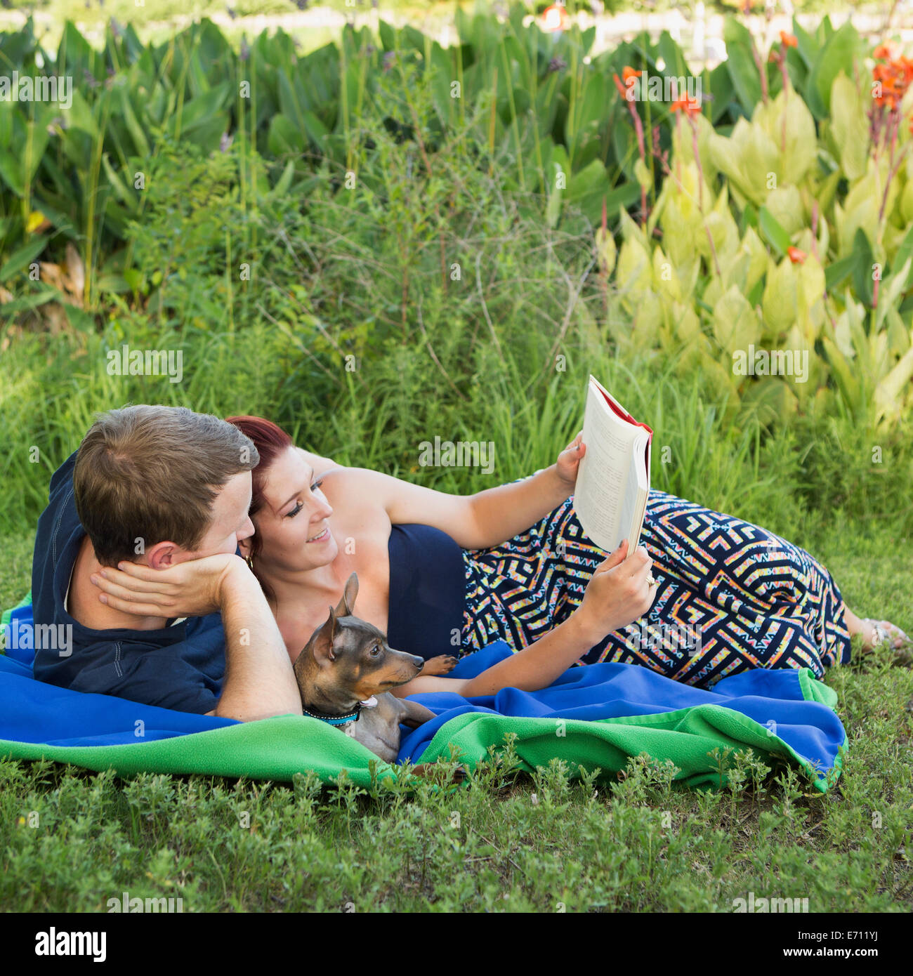 Ein paar, Mann und Frau sitzen auf einer Picknickdecke, ein Buch mit ihrem kleinen Hund zwischen ihnen. Stockfoto