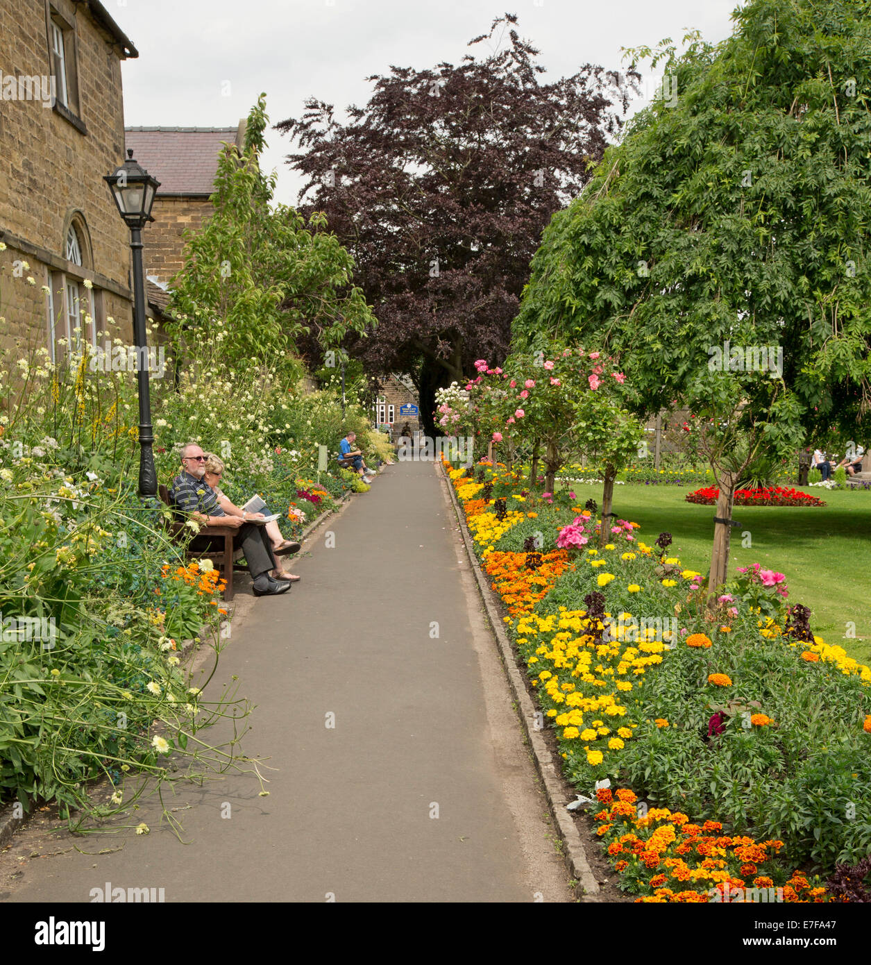 Bunte Anzeige von Frühlingsblumen neben Wiesen und Bäumen mit Menschen entspannen auf Parkbank in Bad Gärten, Bakewell, England Stockfoto