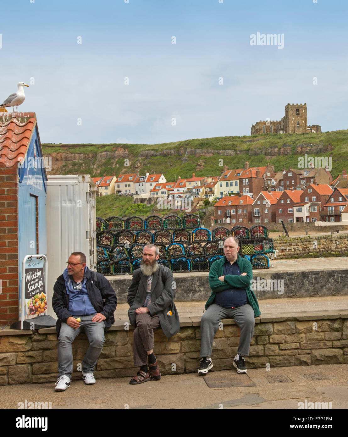 Drei ältere Männer sitzen an Wand neben Hafen an der englischen Küste Stadt Whitby mit Häusern auf Hügel im Hintergrund Stockfoto