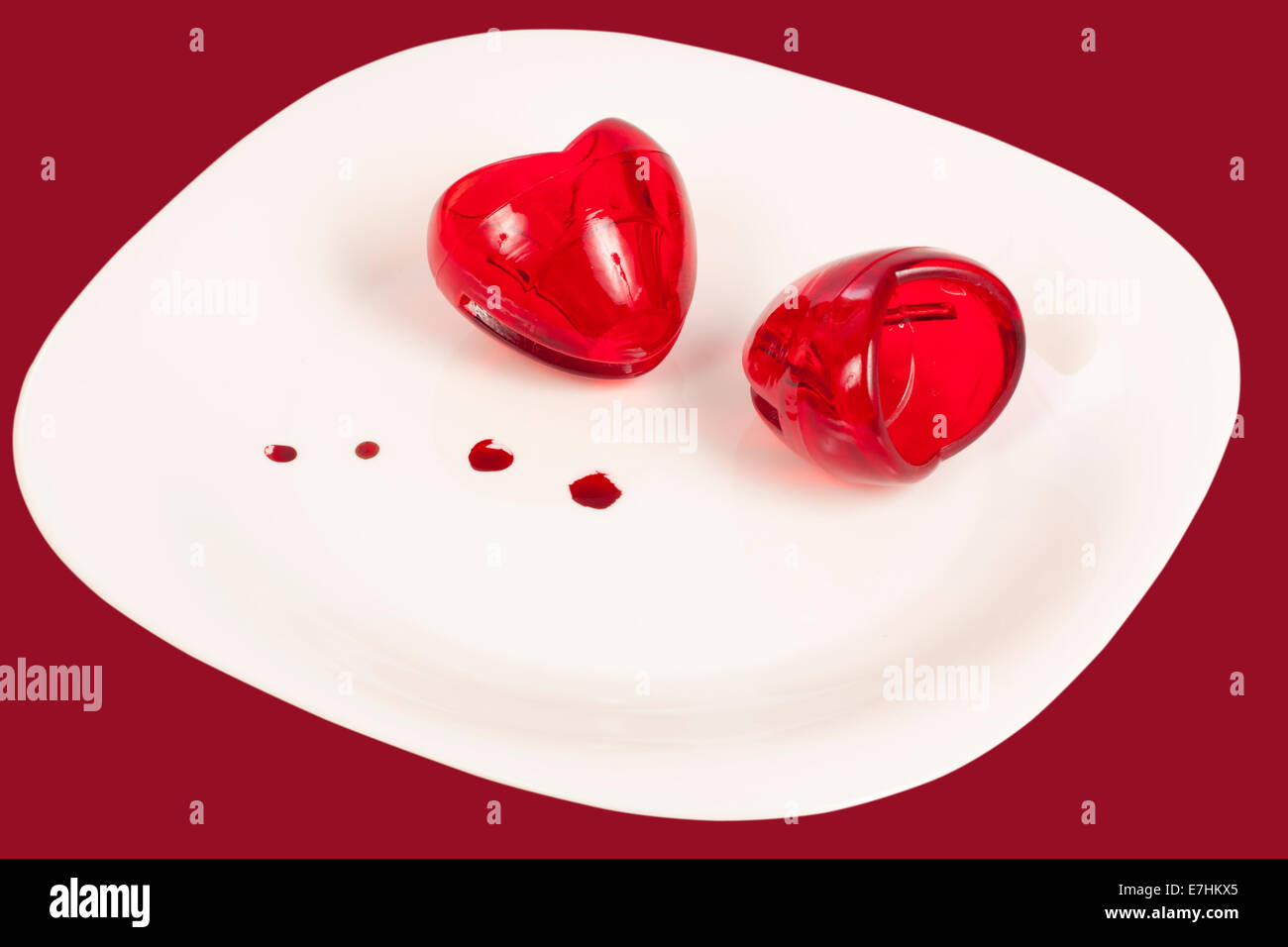 Zwei Herzen aus Glas auf einen Teller geben und ein paar Tropfen Blut isoliert mit Beschneidungspfad auf rotem Grund Stockfoto
