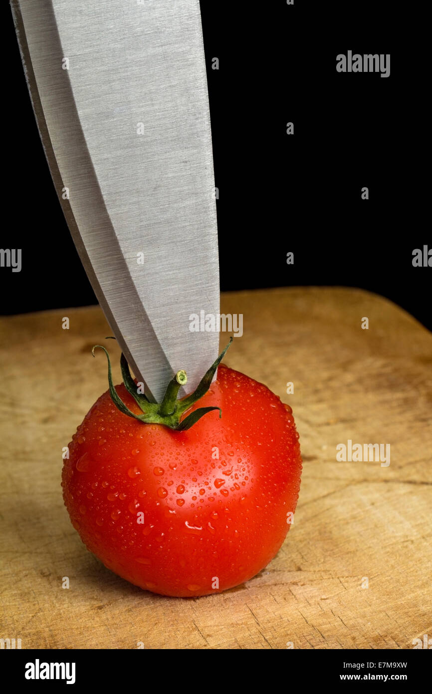 Rote Tomate und ein Stahl Messer auf einem Holzbrett mit schwarzem Hintergrund Stockfoto