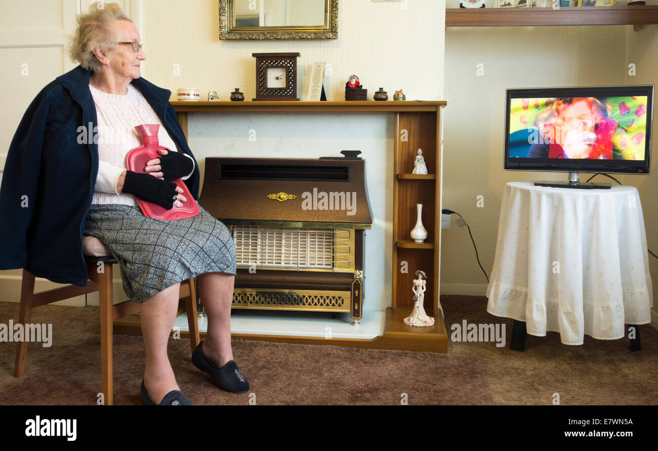 Neunzig Jahre alte Dame, die Handschuhe trägt und eine Wärmflasche in der Hand hält, während sie in ihrem eigenen Haus Fernsehen und dabei das Gasfeuer ausgeschaltet ist. Stockfoto