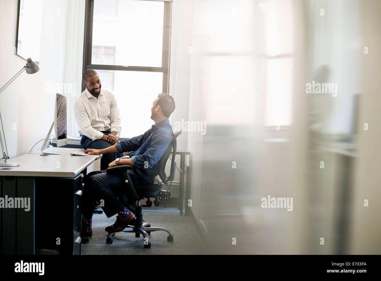 Büroalltag. Ein Mann lehnt sich zurück in einen Bürostuhl im Gespräch mit einem Kollegen sitzt auf dem Rand des Schreibtisches. Stockfoto