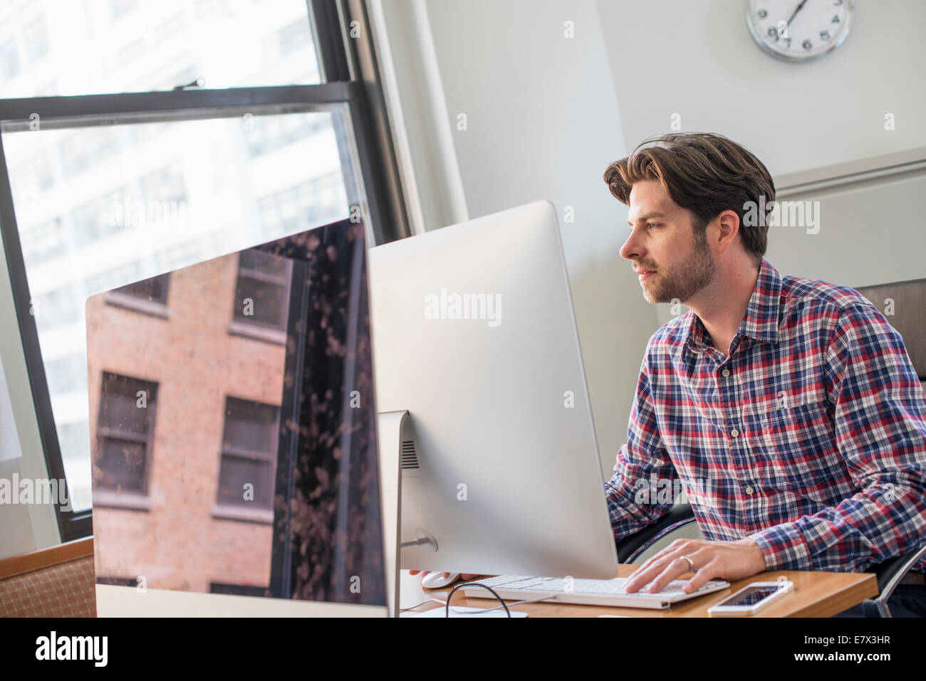 Ein Mann in Achecked Shirt sitzt an einem Schreibtisch mit einem Computer. Stockfoto