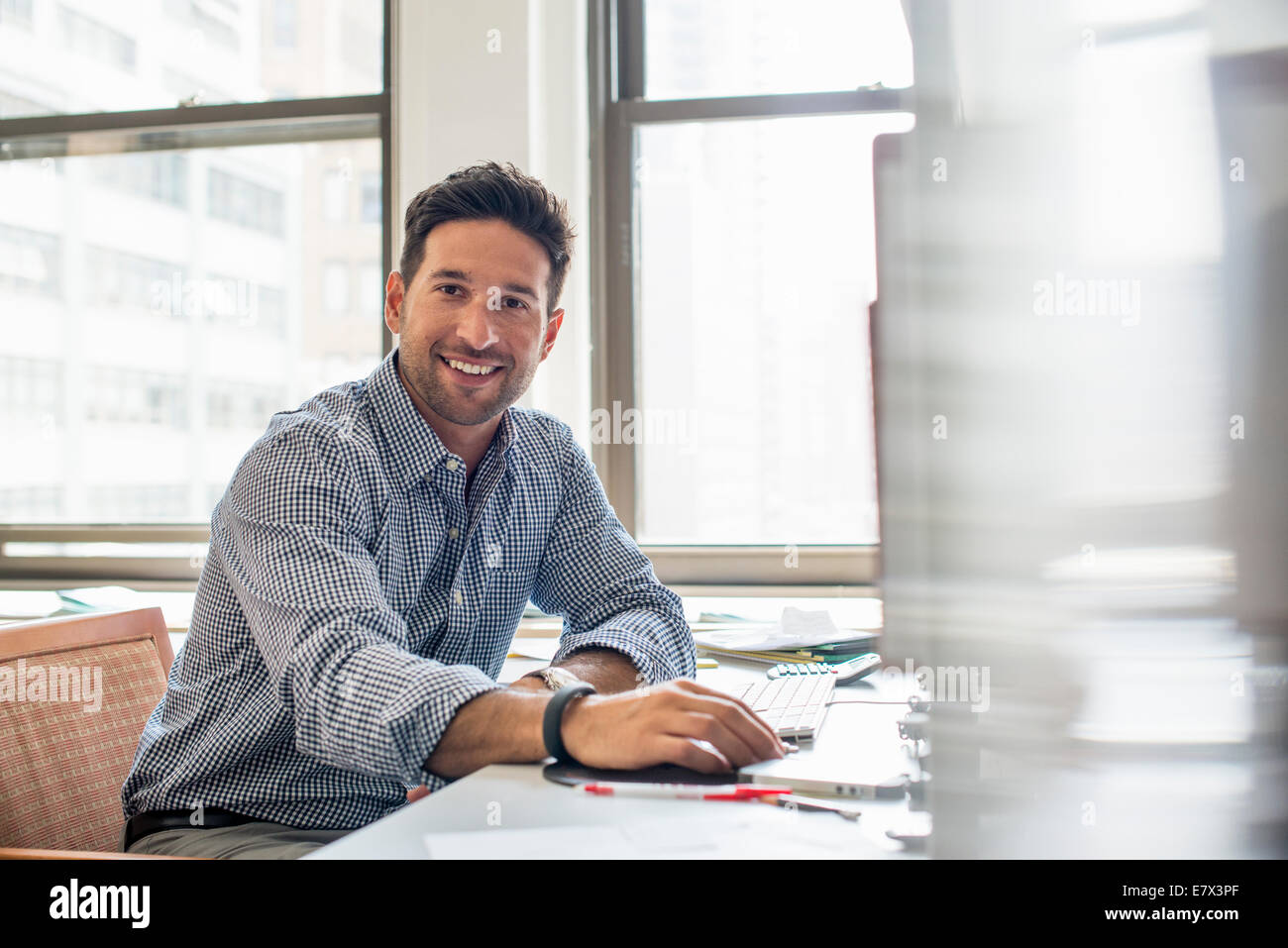 Büroalltag. Ein Mann sitzt an einem Schreibtisch mit einem Computer in die Kamera schaut. Stockfoto