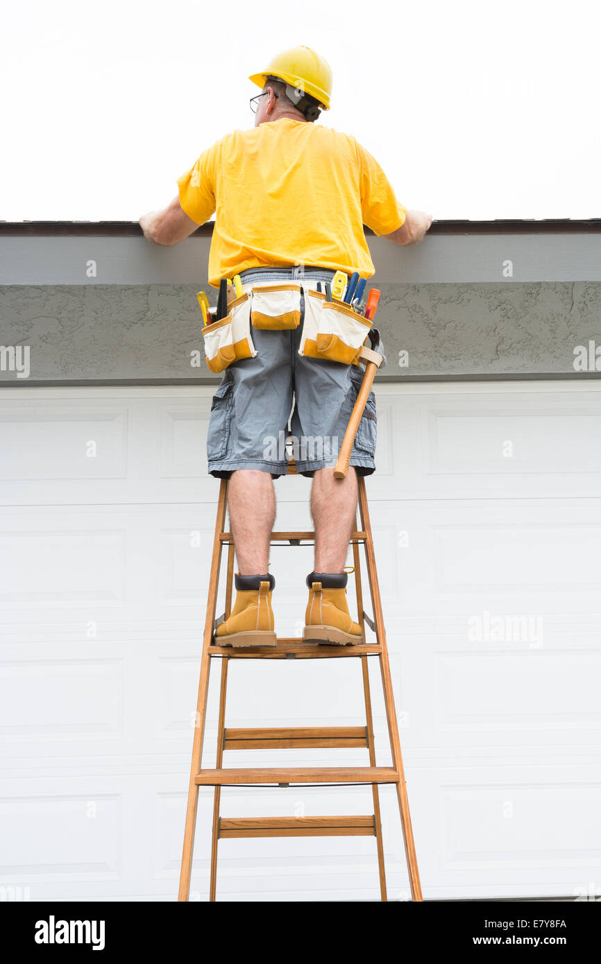 Ein Unternehmer mit einem Gürtel voller Werkzeuge geht auf eine Leiter auf das Dach von einem Heim zu inspizieren. Stockfoto