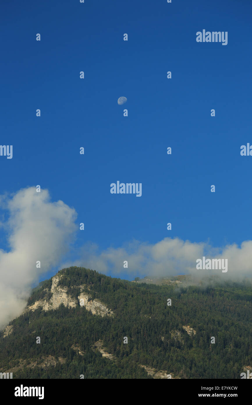 Ein Foto des Mondes in einem blauen Morgenhimmel über einen Berg mit einer Bedeckung von Wolken. Stockfoto