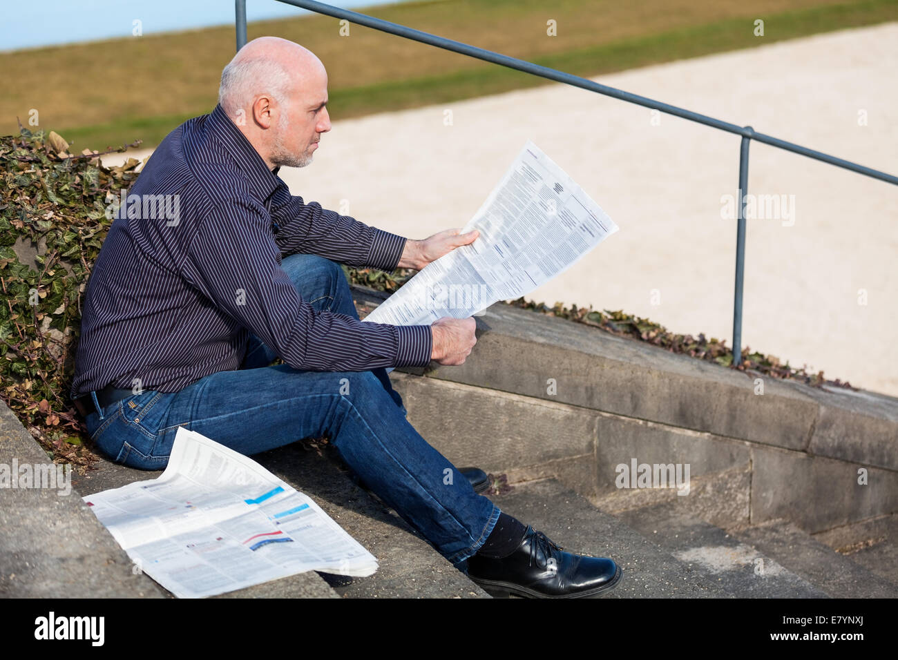 Glatze Mann mittleren Alters in Jeans sitzen im Freien in der Sonne auf einer Treppe eine Zeitung lesen Stockfoto