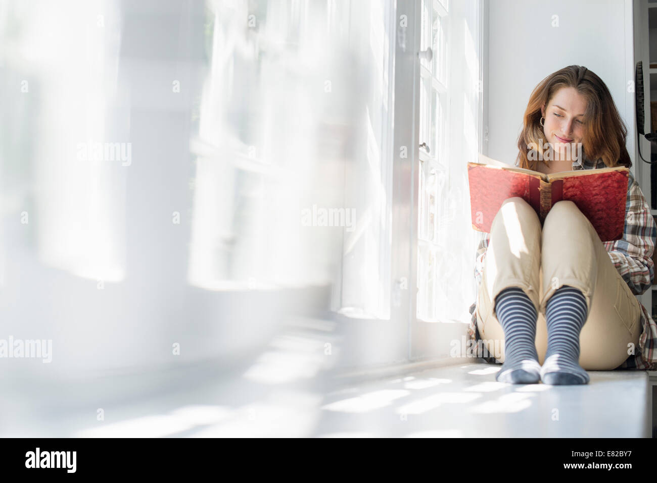 Eine Frau sitzt an einem Fenster lesen mit einem Buch auf dem Schoß. Stockfoto