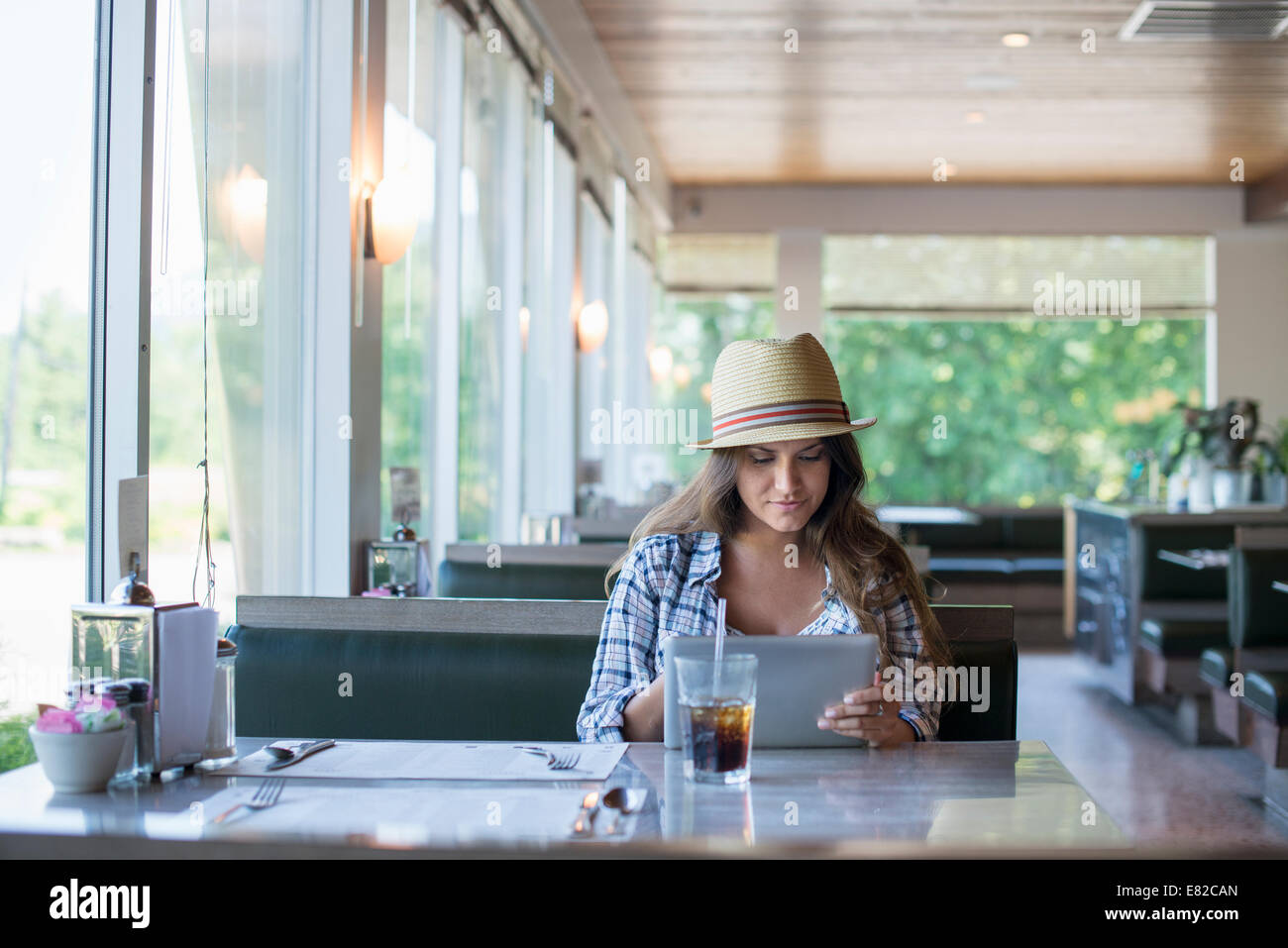 Eine junge Frau sitzt an einem Tisch in einem Diner, trägt einen Strohhut mit einem digitalen Tablet. Stockfoto