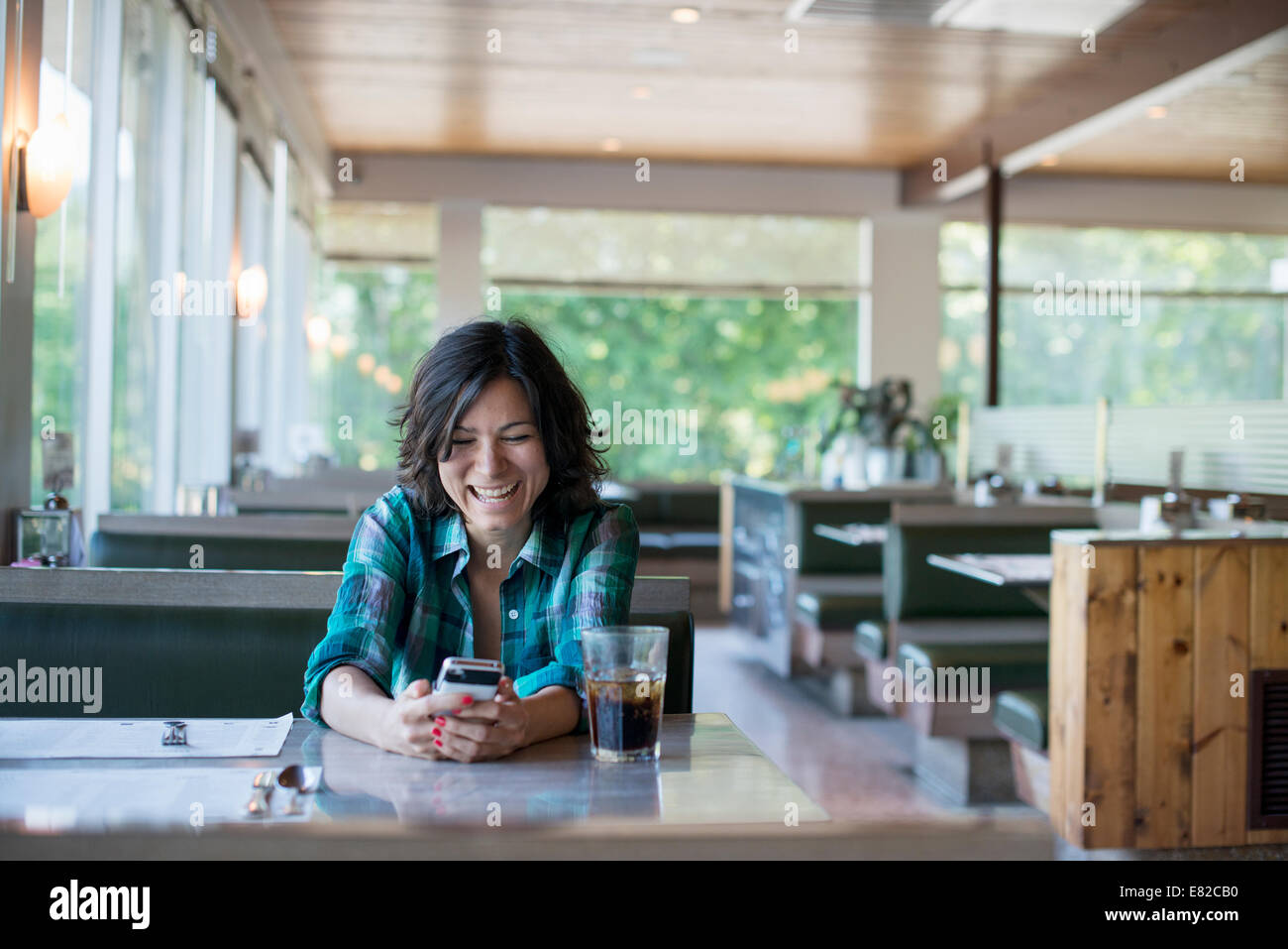 Eine Frau in ein kariertes Hemd an einem Tisch sitzen, lachen und mit Blick auf ihr Smartphone. Stockfoto