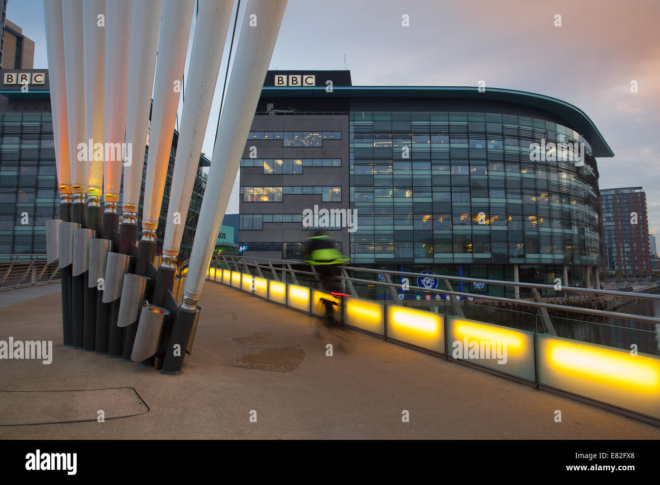 BBC's Flaggschiff-Fernsehzentrale im Norden in MediaCity, Media City, Salford Quays mit einem Jogger, der die MediaCityUK Link Fußgängerbrücke überquert, die im Morgengrauen beleuchtet wird. Stockfoto