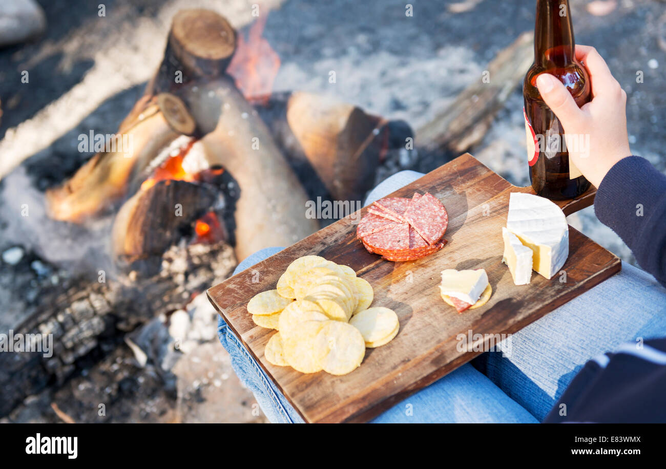 Gourmet-camping Essen auf einem Holzbrett mit Käse, Salami und Cracker mit einem Bier vor dem Kamin Stockfoto