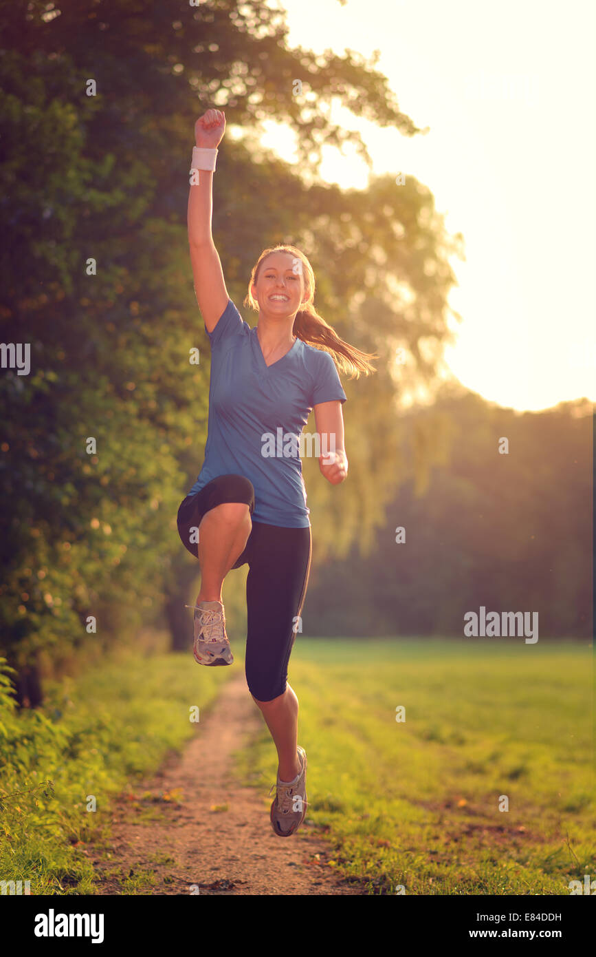 Energische Frau springt in die Luft mit einem glücklichen Lächeln voller Vitalität, wie sie entlang verläuft ein Landes zu verfolgen, während des Trainings Stockfoto