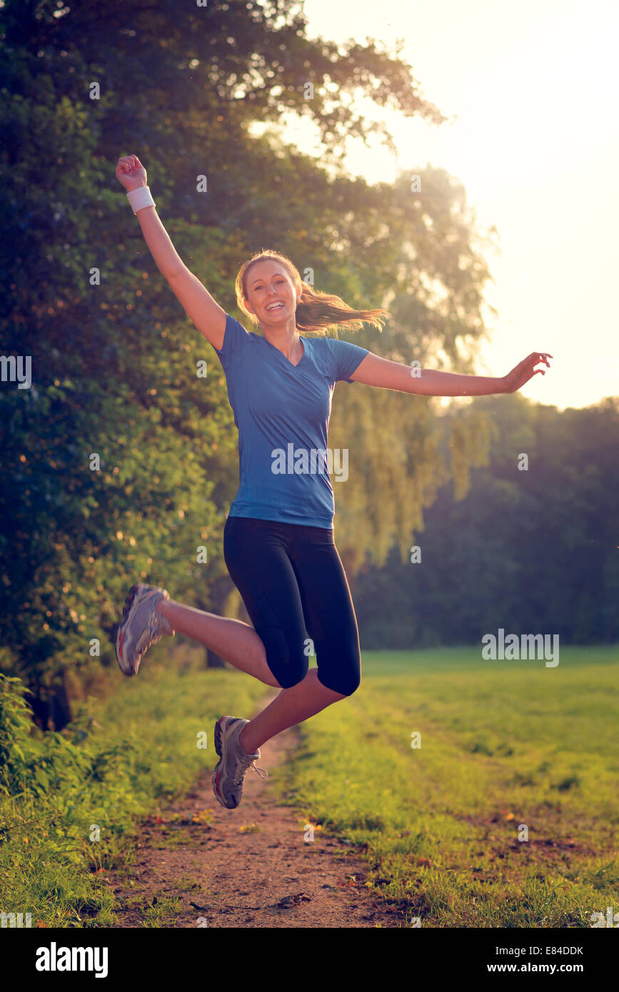 Energische Frau springt in die Luft mit einem glücklichen Lächeln voller Vitalität, wie sie entlang verläuft ein Landes zu verfolgen, während des Trainings Stockfoto