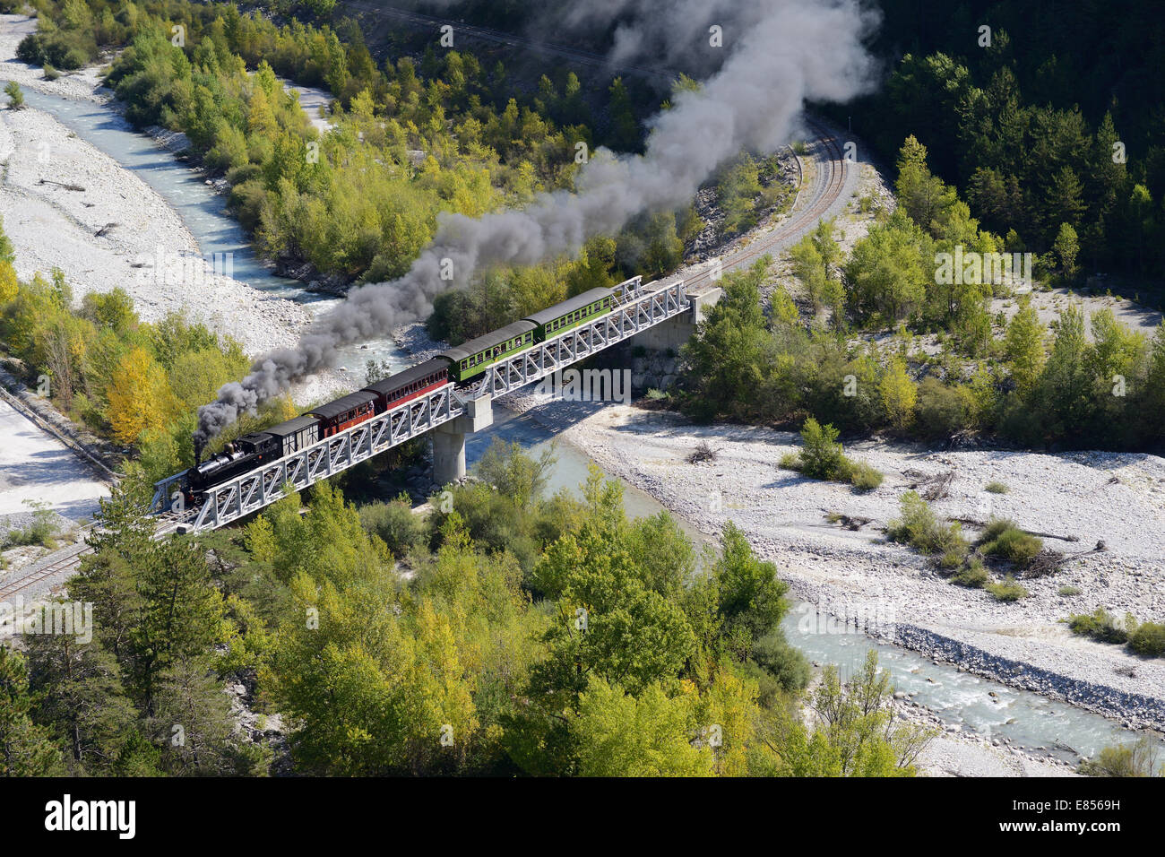 LUFTAUFNAHME. Historische und touristische Dampfeisenbahn auf einer Schotterbrücke über den Fluss Var. Train des Pignes zwischen Entrevaux und Annot, Frankreich. Stockfoto