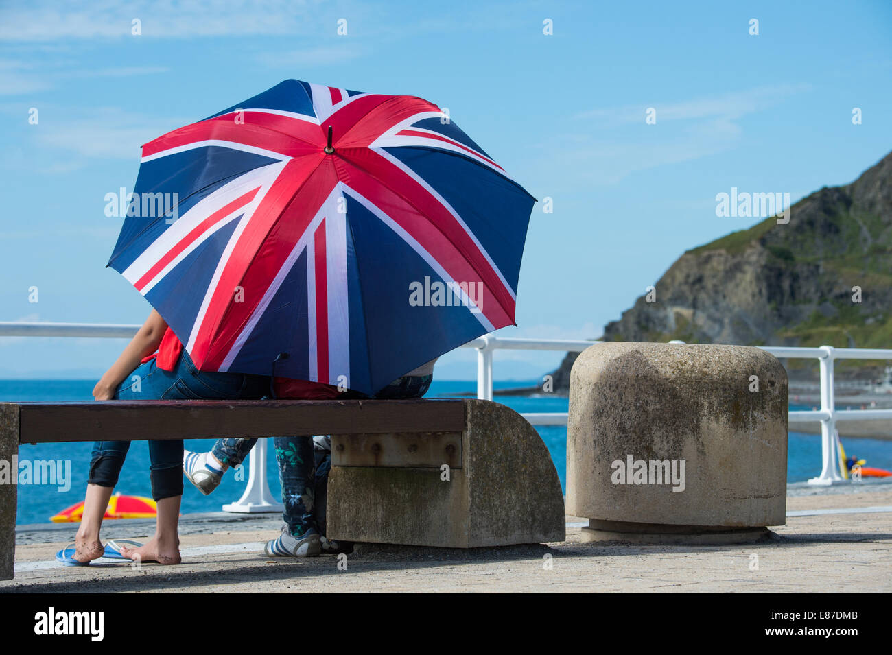 Eine oder mehrere Personen sitzen auf Bank bergende unter Gösch Regenschirm Brolly am Meer auf heißen Juli Sommer Nachmittag UK Stockfoto