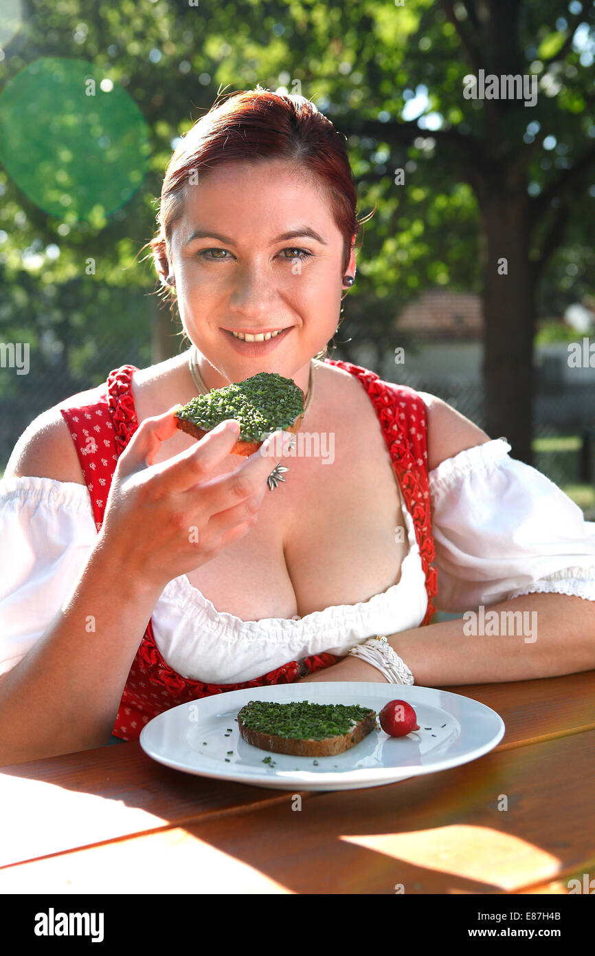 Rote Kopf Frau in typisch bayrischer Tracht isst Brot mit Schnittlauch in einem bayerischen Biergarten Stockfoto