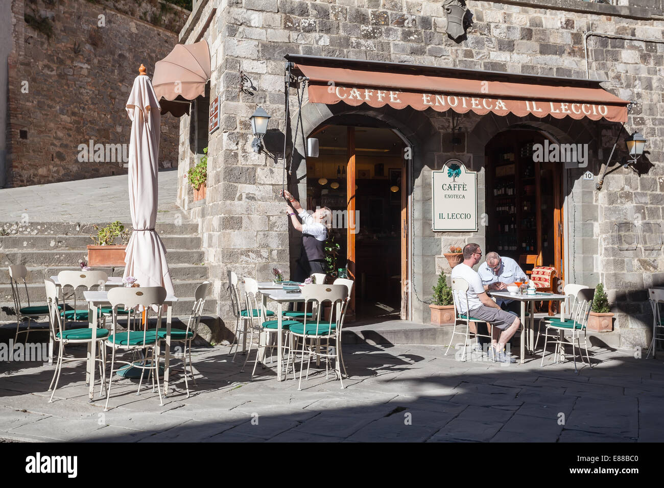 Zwei Männer draußen auf eine Café-Bar-Terrasse "Il Leccio"; Frau-Barkeeper, die gerade geöffnet und versucht, die Abdeckung Banner roll Stockfoto