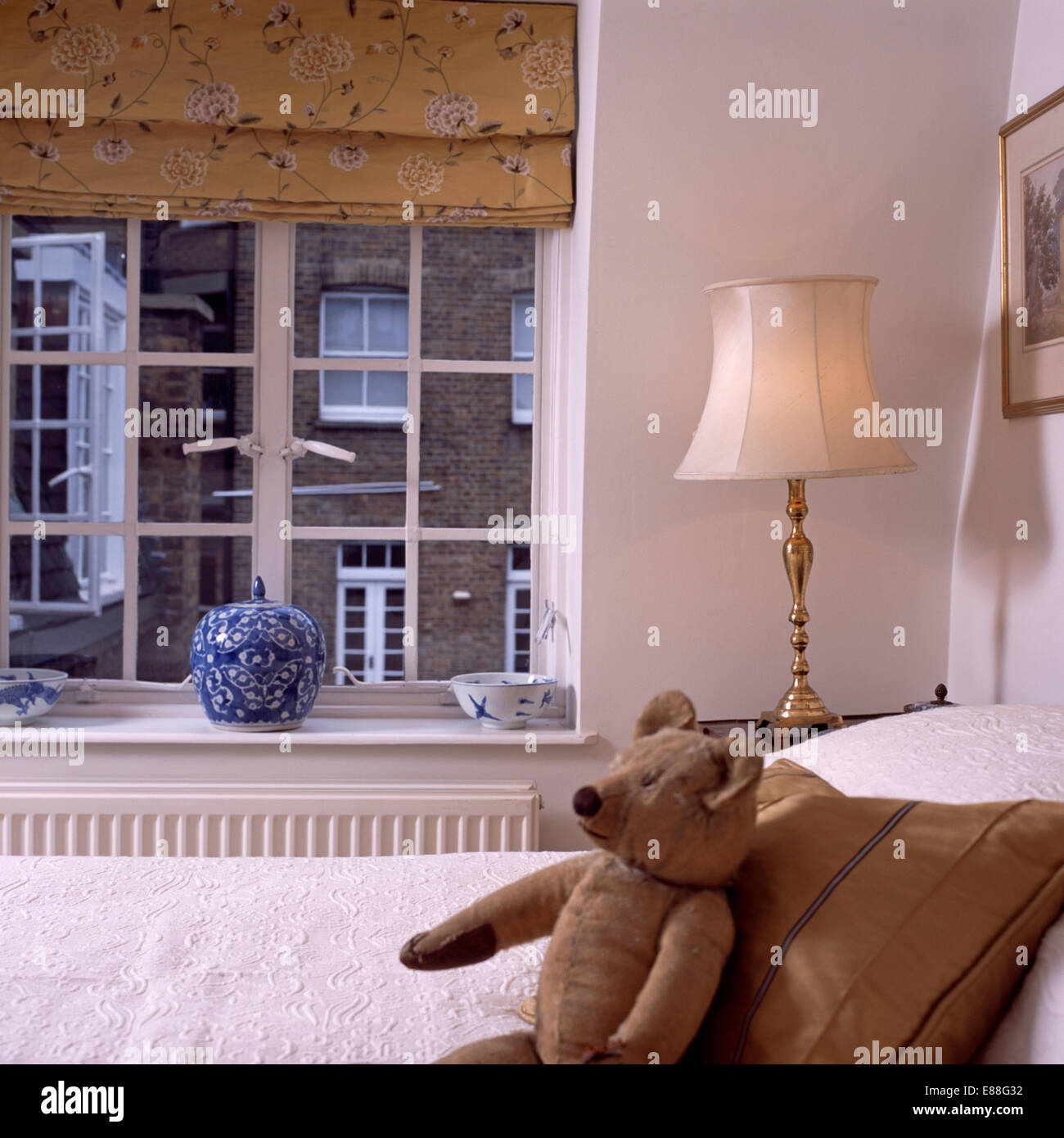 Lampe neben Bett mit alten Teddybär und Seide Kissen vor Fenster mit gemusterten neutral blind Stockfoto