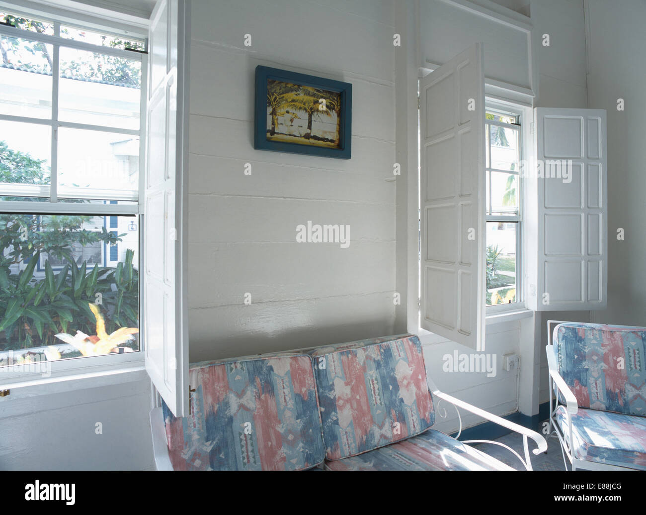 Weißen Fensterläden aus Holz unter Windows in weiße Küste Schlafzimmer mit gemusterten Kissen auf Metallstühlen Stockfoto