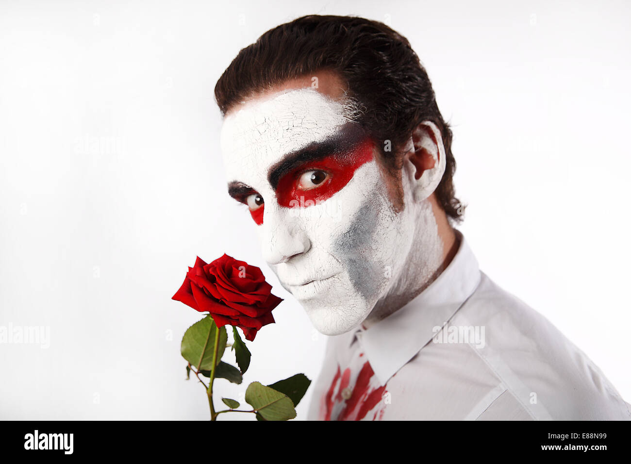 Mann mit weißen Mascara und blutigen Shirt hält rote Rose vor einem weißen Hintergrund Stockfoto