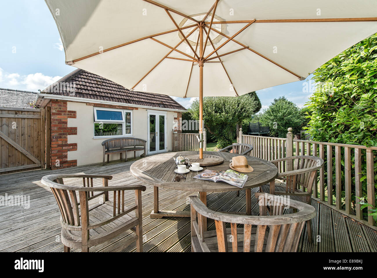 Ein großer Sonnenschirm für Nachmittagstee serviert auf einem hölzernen Gartentisch & Möbel. außen am sonnigen Tag. UK Stockfoto