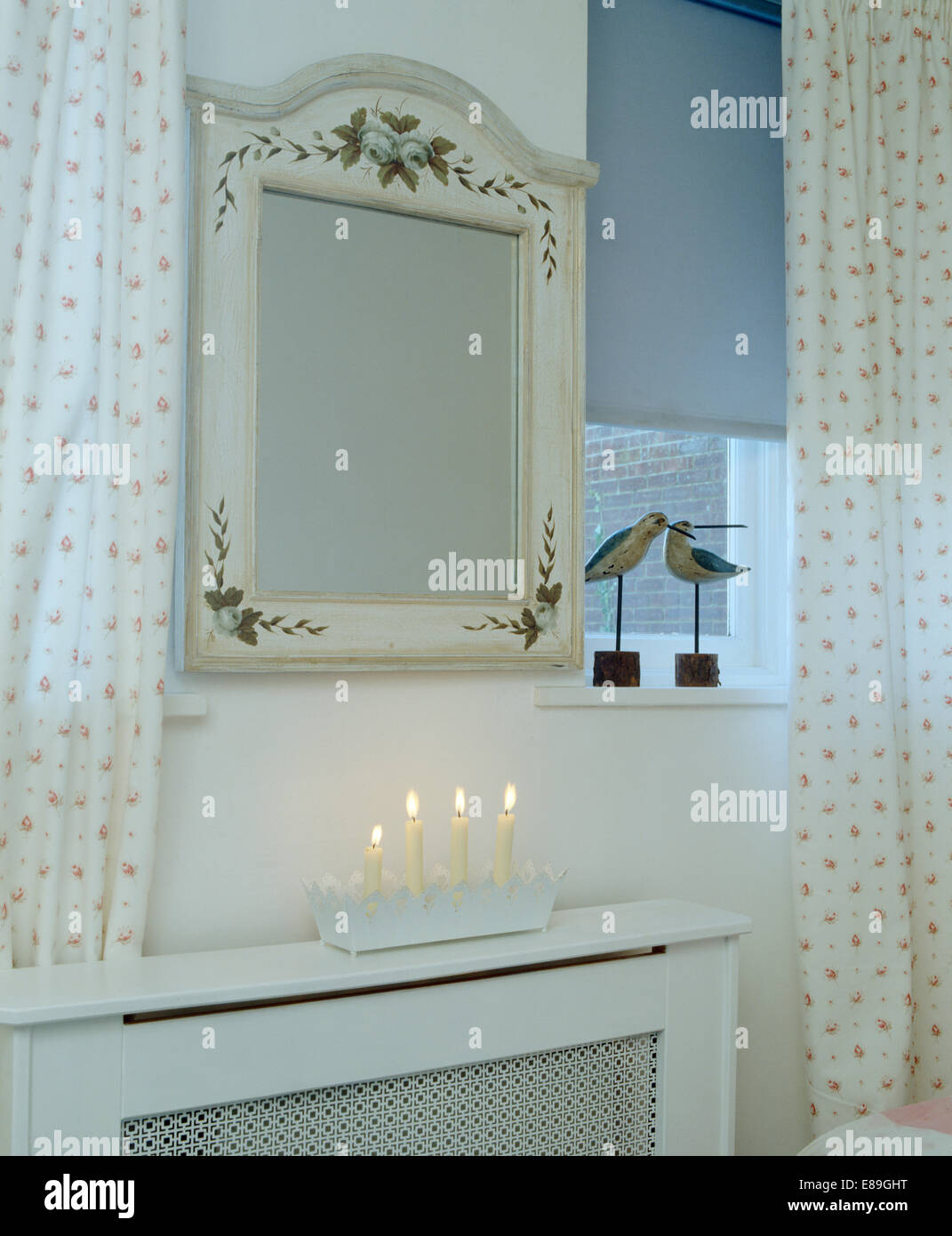 Blaue blind und gemusterte Vorhänge neben Hand gemalte Spiegel über Heizkörper Abdeckung mit brennenden Kerzen Stockfoto
