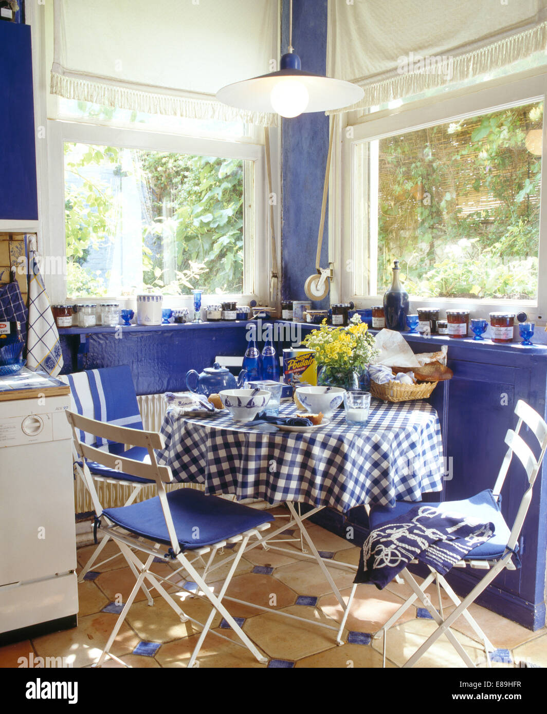 Weiße Klappstühle an kleinen runden Tisch mit blau + weiß aufgegebenes Tuch vor Fenstern mit weißen blind Stockfoto